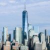 「ニューヨーク市のワン・ワールド・トレード・センターが中心に見えるスカイライン。川から見た青空の下、ガラスとスチールの高層ビルが連なる都市景観。」