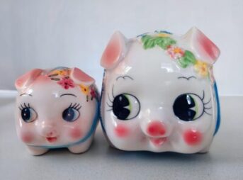 「花柄が描かれた二つの陶器製の豚の貯金箱。一方は顔を正面に向け、もう一方が小さくもう一方が一回り大きくまるで、親子みたいです。両方とも大きな瞳とピンクのほっぺたが特徴的です。」